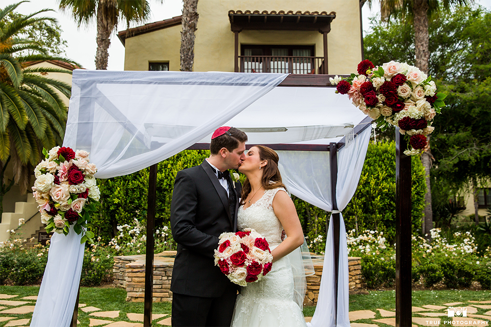 San diego beach wedding at estancia la jolla bride form fitting strapless gown with sweetheart neckline and lace design with long veil and groom black shawl lapel tuxedo with matching vest and white dress shirt with black bow tie and white pocket square with white floral boutonniere kissing under altar during ceremony