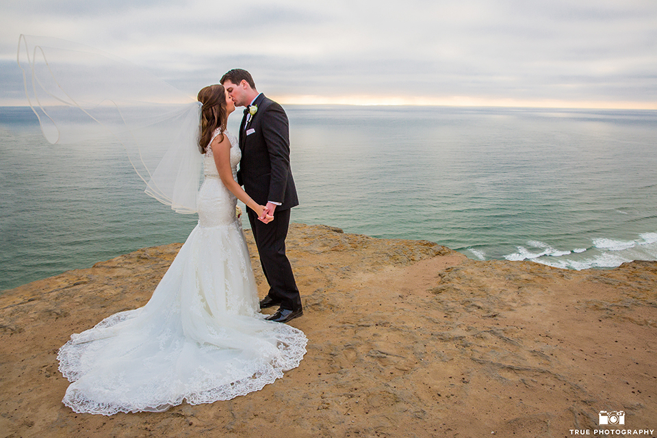 San diego beach wedding at estancia la jolla bride form fitting strapless gown with sweetheart neckline and lace design with long veil and groom black shawl lapel tuxedo with matching vest and white dress shirt with black bow tie and white pocket square with white floral boutonniere holding hands and kissing