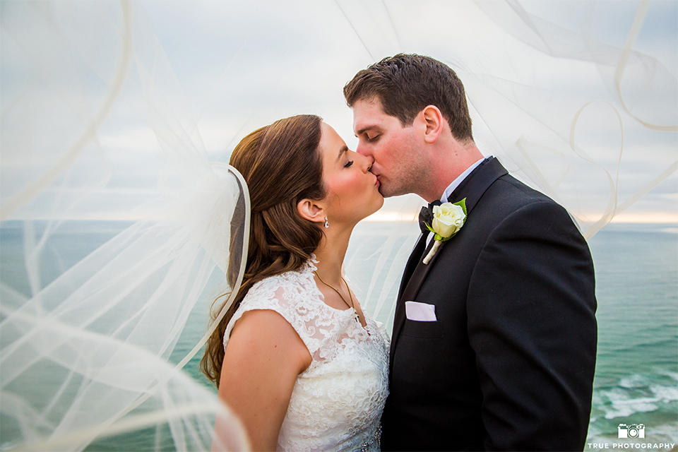 San diego beach wedding at estancia la jolla bride form fitting strapless gown with sweetheart neckline and lace design with long veil and groom black shawl lapel tuxedo with matching vest and white dress shirt with black bow tie and white pocket square with white floral boutonniere kissing under veil close up