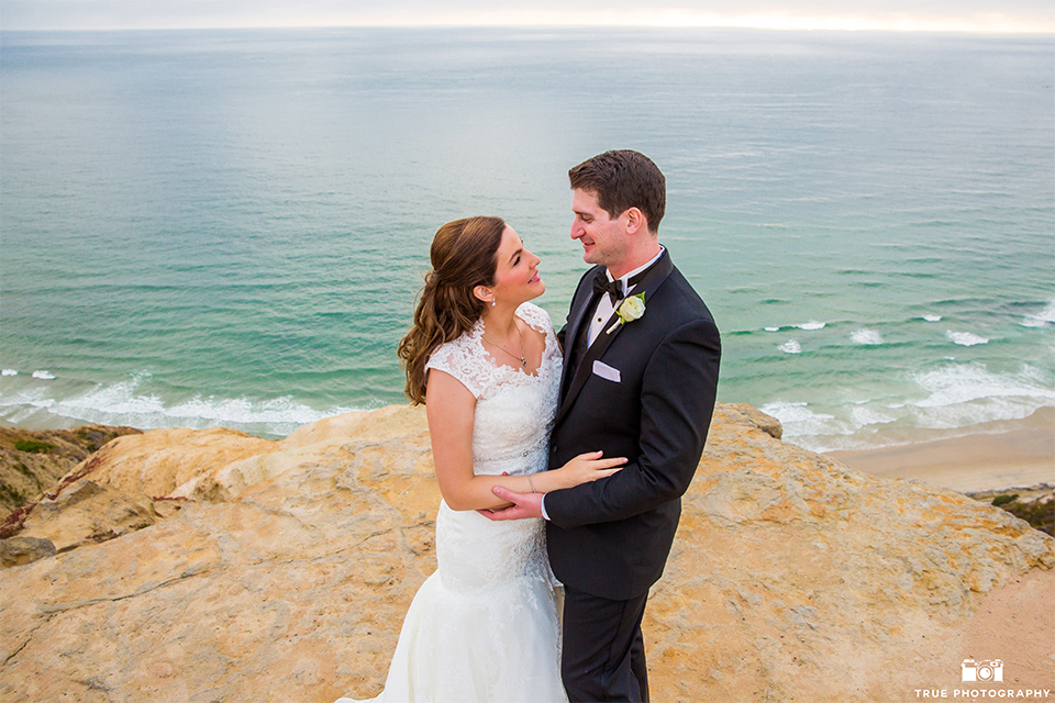 San diego beach wedding at estancia la jolla bride form fitting strapless gown with sweetheart neckline and lace design with long veil and groom black shawl lapel tuxedo with matching vest and white dress shirt with black bow tie and white pocket square with white floral boutonniere hugging