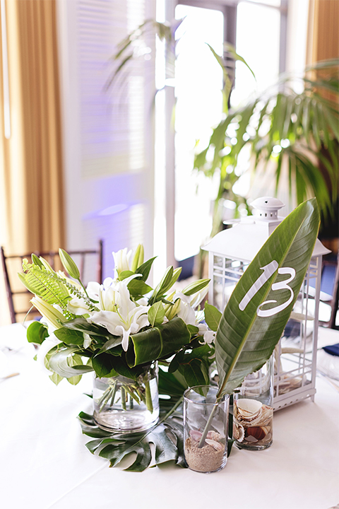 Huntington beach wedding at the hilton waterfront resort table set up white table linen with white and green floral centerpiece decor with white table numbers written on green leaves and glass candle decor with dark chairs