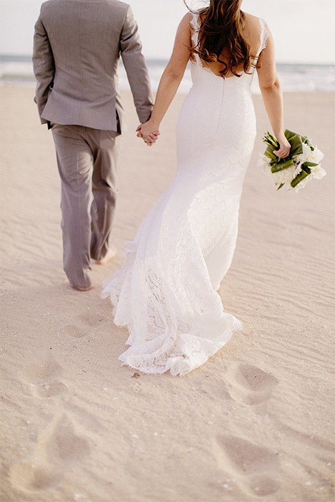 Huntington beach wedding at the hilton waterfront resort bride form fitting lace gown with a plunging neckline and thin straps with lace detail and low back design with groom heather grey notch lapel suit with matching vest and white dress shirt with long navy blue patterned tie and white floral boutonniere holding hands and bride holding white and green floral bridal bouquet