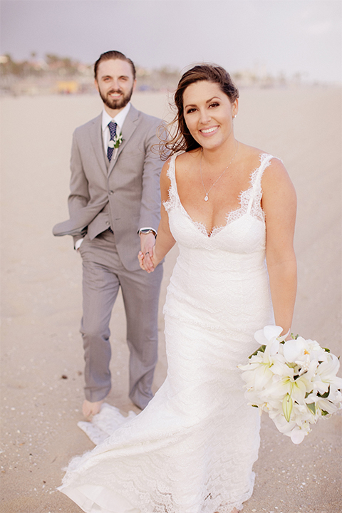 Huntington beach wedding at the hilton waterfront resort bride form fitting lace gown with a plunging neckline and thin straps with lace detail and low back design with groom heather grey notch lapel suit with matching vest and white dress shirt with long navy blue patterned tie and white floral boutonniere holding hands and walking bride holding white and green floral bridal boquuet