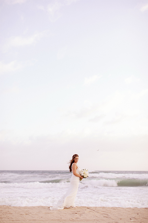 Huntington beach wedding at the hilton waterfront resort bride form fitting lace gown with a plunging neckline and thin straps with lace detail and low back design holding white and green floral bridal bouquet on the beach