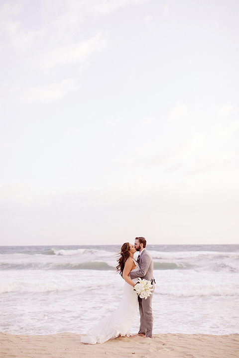 Huntington beach wedding at the hilton waterfront resort bride form fitting lace gown with a plunging neckline and thin straps with lace detail and low back design with groom heather grey notch lapel suit with matching vest and white dress shirt with long navy blue patterned tie and white floral boutonniere standing and kissing bride holding white and green floral bridal bouquet