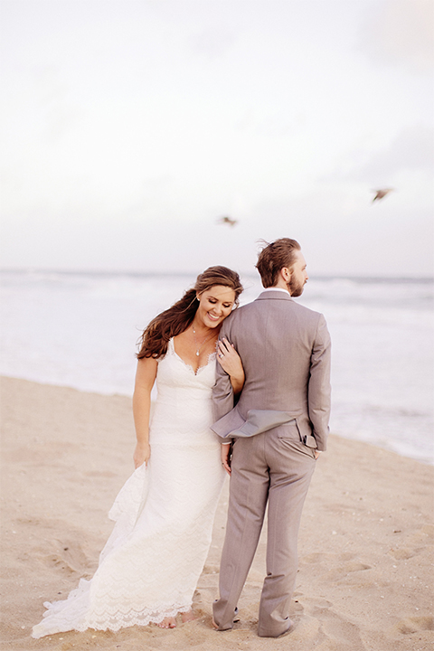 Huntington beach wedding at the hilton waterfront resort bride form fitting lace gown with a plunging neckline and thin straps with lace detail and low back design with groom heather grey notch lapel suit with matching vest and white dress shirt with long navy blue patterned tie and white floral boutonniere standing and hugging arm bride holding white and green floral bridal bouquet