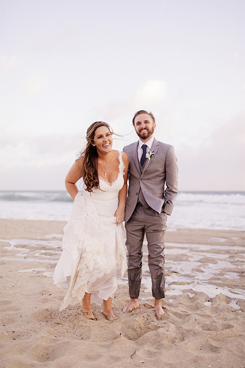 Huntington beach wedding at the hilton waterfront resort bride form fitting lace gown with a plunging neckline and thin straps with lace detail and low back design with groom heather grey notch lapel suit with matching vest and white dress shirt with long navy blue patterned tie and white floral boutonniere standing and holding hands on the beach