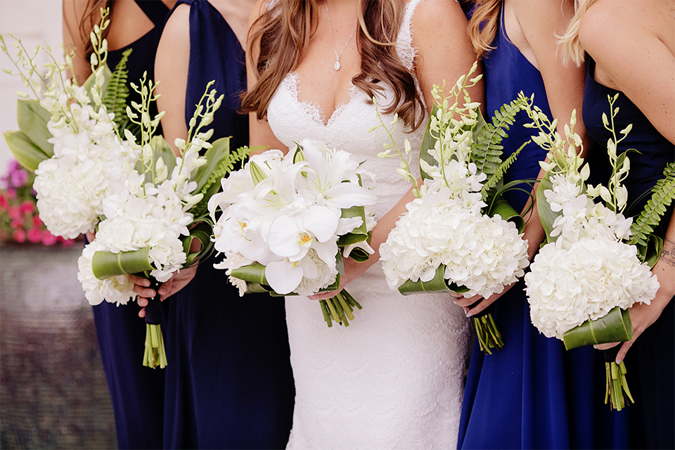 Huntington beach wedding at the hilton waterfront resort bride form fitting lace gown with a plunging neckline and thin straps with lace detail and low back design holding white and green floral bridal bouquet with bridesmaids long navy blue dresses with white and green floral bouquets close up