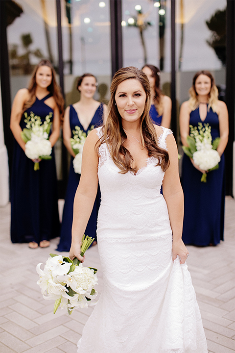 Huntington beach wedding at the hilton waterfront resort bride form fitting lace gown with a plunging neckline and thin straps with lace detail and low back design holding white and green floral bridal bouquet with bridesmaids long navy blue dresses holding white and green floral bouquets