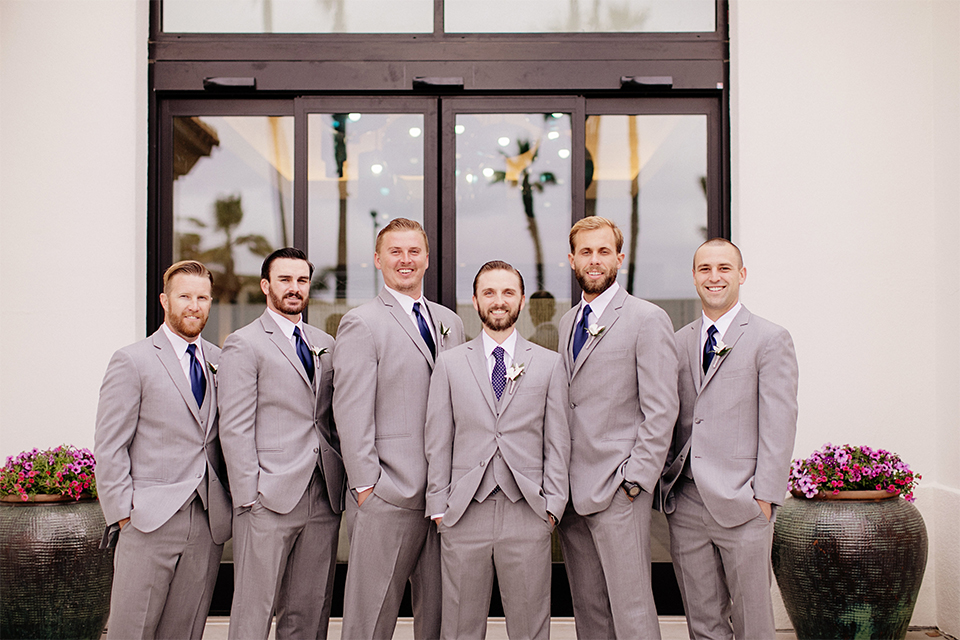 Huntington beach wedding at the hilton waterfront resort groom and groomsmen heather grey notch lapel suits with matching vests and white dress shirt with long navy blue patterned tie and white floral boutonnieres standing with hands in pockets