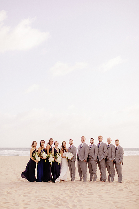 Huntington beach wedding at the hilton waterfront resort bride form fitting lace gown with a plunging neckline and thin straps with lace detail and low back design with groom heather grey notch lapel suit with matching vest and white dress shirt with long navy blue patterned tie and white floral boutonniere with wedding party bridesmaids long navy dresses with white floral bouquets and groomsmen heather grey suits with long navy blue ties