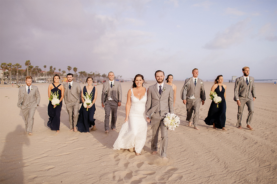 Huntington beach wedding at the hilton waterfront resort bride form fitting lace gown with a plunging neckline and thin straps with lace detail and low back design with groom heather grey notch lapel suit with matching vest and white dress shirt with long navy blue patterned tie and white floral boutonniere walking with wedding party on the beach bridesmaids long navy blue dresses with white floral bouquets and groomsmen heather grey suits with long navy blue ties