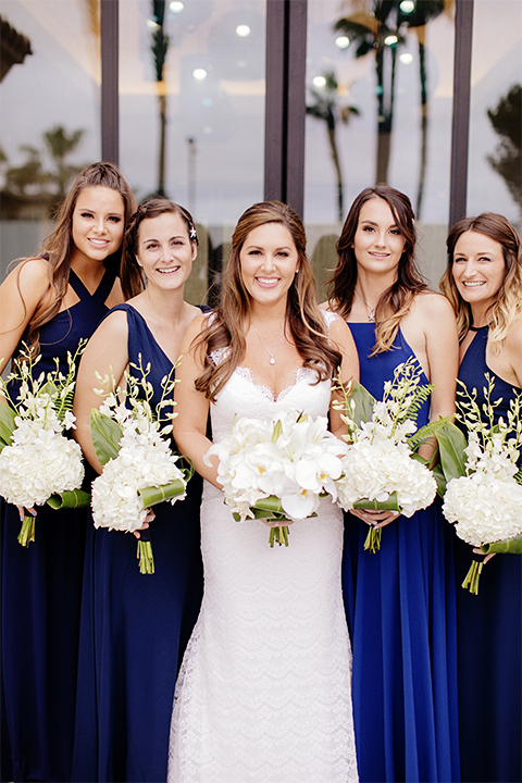 Huntington beach wedding at the hilton waterfront resort bride form fitting lace gown with a plunging neckline and thin straps with lace detail and low back design holding white and green floral bridal bouquet with bridesmaids long navy blue dresses holding white and green floral bouquets