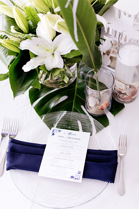 Huntington beach wedding at the hilton waterfront resort table set up white table linen with white and green floral centerpiece decor with white table numbers written on green leaves and glass candle decor with dark chairs and navy napkin decor