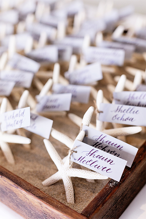 Huntington beach wedding at the hilton waterfront resort table set up white table linen with white and green floral centerpiece decor with white table numbers written on green leaves and glass candle decor with dark chairs with white sea shells and guests names