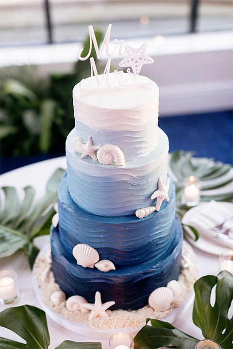 Huntington beach wedding at the hilton waterfront resort table set up white table linen with white and green floral centerpiece decor with white table numbers written on green leaves and glass candle decor with dark chairs wedding cake five tier blue and white ombre cake with white sea shell decor and green leaves on table