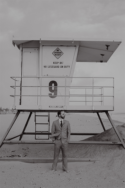 Huntington beach wedding at the hilton waterfront resort groom heather grey notch lapel suit with matching vest and white dress shirt with long navy patterned tie and white floral boutonniere standing by lifeguard tower on beach black and white photo