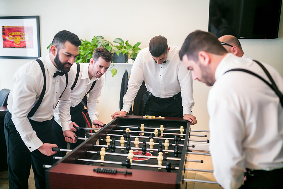 San juan capistrano outdoor wedding at plaza de magdelena groom black notch lapel tuxedo with white dress shrirt and black bow tie with white floral boutonniere playing foosball with groomsmen getting ready before wedding black tuxedos with black bow tie and black suspenders