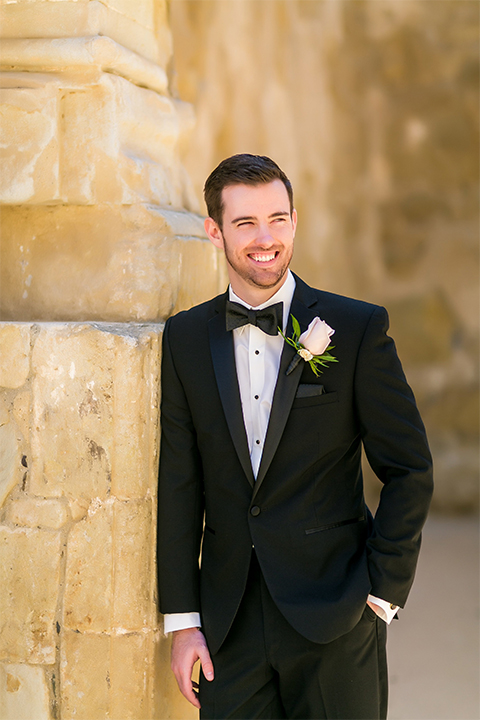 San juan capistrano outdoor wedding at plaza de magdelena groom black notch lapel tuxedo with white dress shrirt and black bow tie with white floral boutonniere standing with hands in pockets
