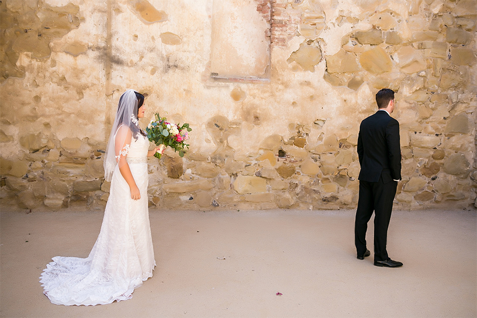 San juan capistrano outdoor wedding at plaza de magdelena bride form fitting gown with lace detail on bodice and thin straps with crystal belt and groom black notch lapel tuxedo with white dress shirt and black bow tie and white floral boutonniere first look