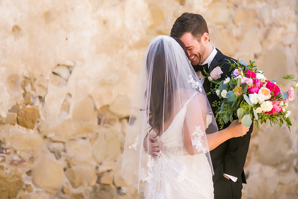 San juan capistrano outdoor wedding at plaza de magdelena bride form fitting gown with lace detail on bodice and thin straps with crystal belt and groom black notch lapel tuxedo with white dress shirt and black bow tie and white floral boutonniere first look bride and groom hugging bride holding white and red floral bridal bouquet