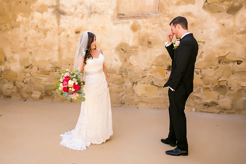 San juan capistrano outdoor wedding at plaza de magdelena bride form fitting gown with lace detail on bodice and thin straps with crystal belt and groom black notch lapel tuxedo with white dress shirt and black bow tie and white floral boutonniere first look bride holding white and red floral bridal bouquet