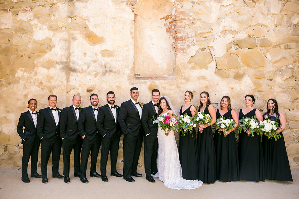 San juan capistrano outdoor wedding at plaza de magdelena bride form fitting gown with lace detail on bodice and thin straps with crystal belt and groom black notch lapel tuxedo with white dress shirt and black bow tie and white floral boutonniere with wedding party bridesmaids long black dresses and groomsmen black tuxedos with black bow ties