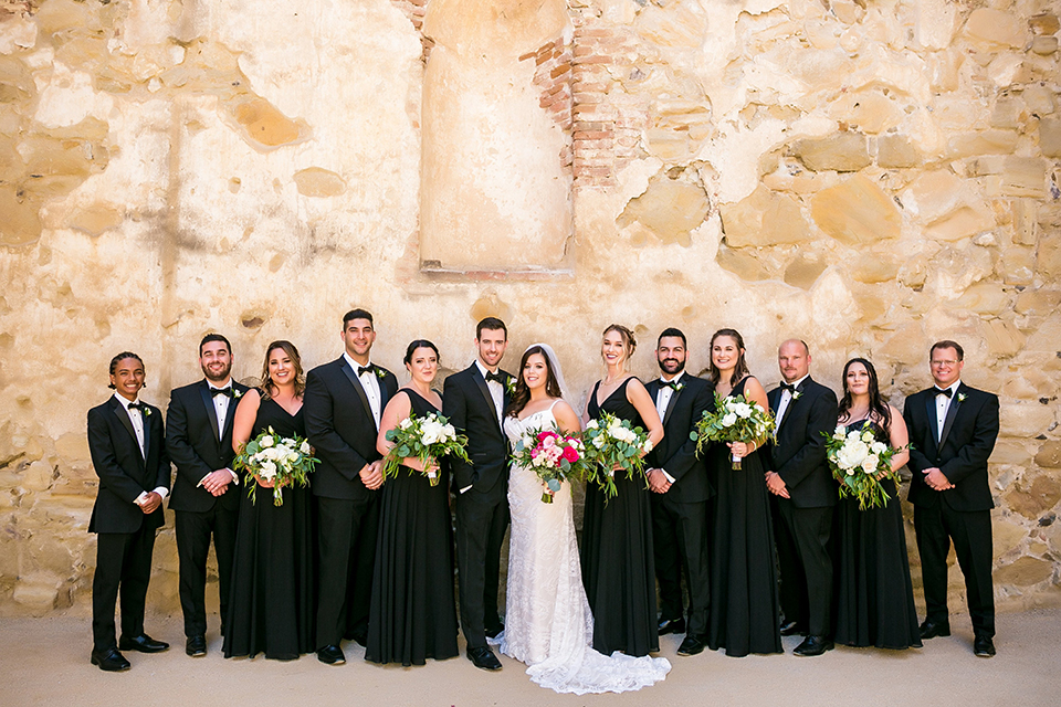 San juan capistrano outdoor wedding at plaza de magdelena bride form fitting gown with lace detail on bodice and thin straps with crystal belt and groom black notch lapel tuxedo with white dress shirt and black bow tie and white floral boutonniere with wedding party bridesmaids long black dresses and groomsmen black tuxedos with black bow ties