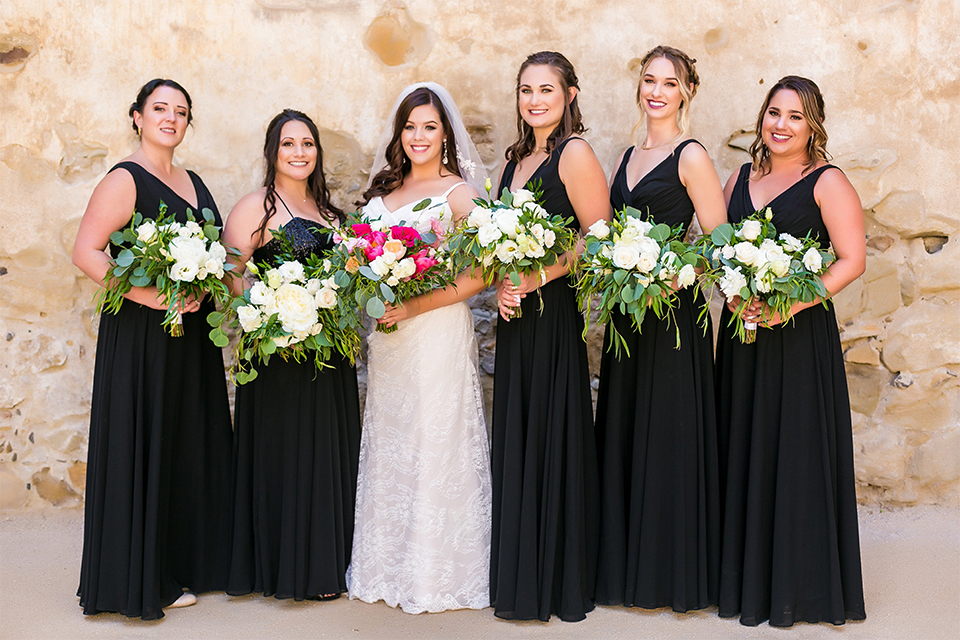 San juan capistrano outdoor wedding at plaza de magdelena bride form fitting gown with lace detail on bodice and thin straps with crystal belt with bridesmaids long black dresses holding white and green floral bridal bouquets