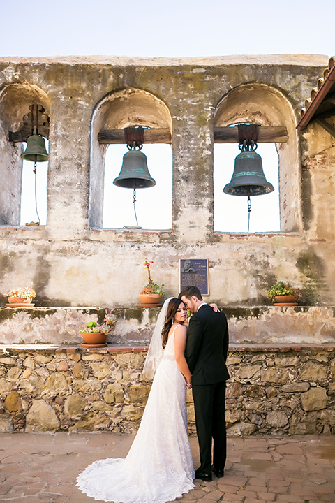 San juan capistrano outdoor wedding at plaza de magdelena bride form fitting gown with lace detail on bodice and thin straps with crystal belt and groom black notch lapel tuxedo with white dress shirt and black bow tie and white floral boutonniere hugging and holding hands