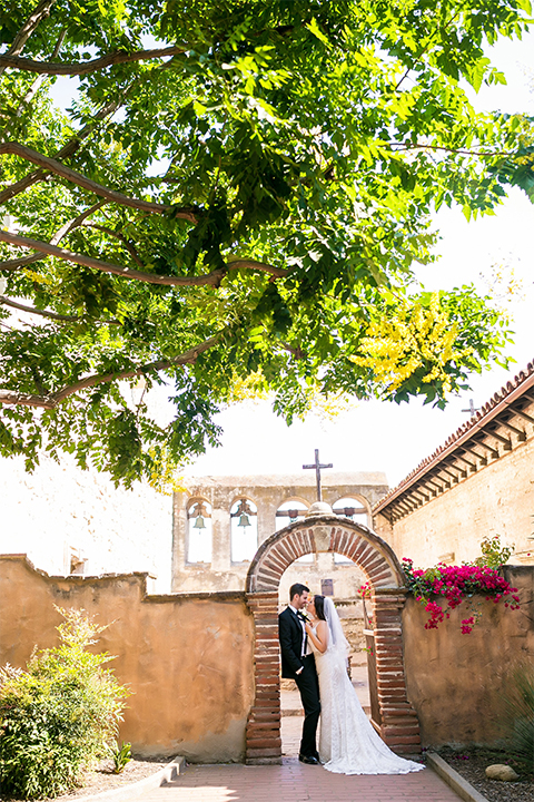 San juan capistrano outdoor wedding at plaza de magdelena bride form fitting gown with lace detail on bodice and thin straps with crystal belt and groom black notch lapel tuxedo with white dress shirt and black bow tie and white floral boutonniere hugging