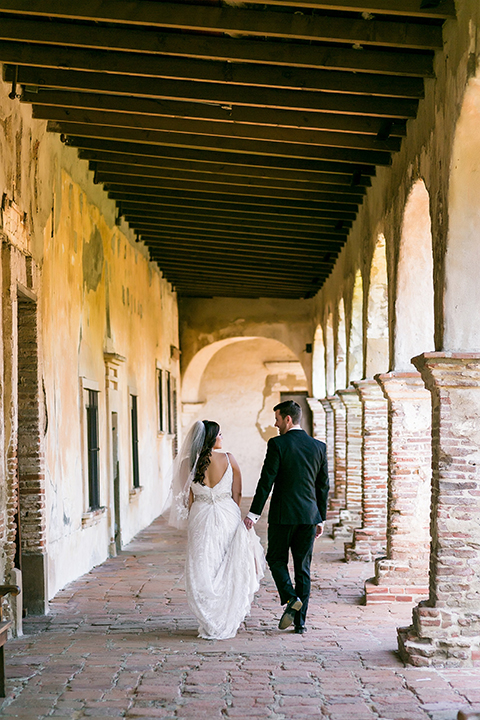 San juan capistrano outdoor wedding at plaza de magdelena bride form fitting gown with lace detail on bodice and thin straps with crystal belt and groom black notch lapel tuxedo with white dress shirt and black bow tie and white floral boutonniere walking and holding hands