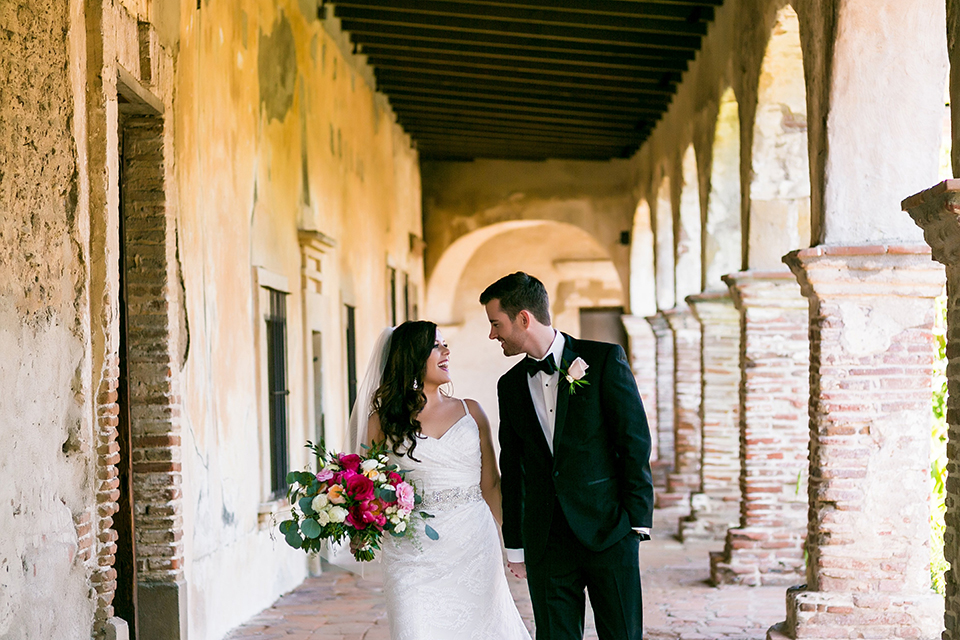 San juan capistrano outdoor wedding at plaza de magdelena bride form fitting gown with lace detail on bodice and thin straps with crystal belt and groom black notch lapel tuxedo with white dress shirt and black bow tie and white floral boutonniere walking and holding hands bride holding white and red floral bridal bouquet
