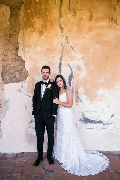San juan capistrano outdoor wedding at plaza de magdelena bride form fitting gown with lace detail on bodice and thin straps with crystal belt and groom black notch lapel tuxedo with white dress shirt and black bow tie and white floral boutonniere hugging