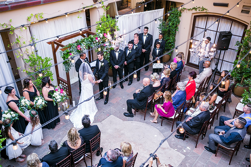 San juan capistrano outdoor wedding at plaza de magdelena bride form fitting gown with lace detail on bodice and thin straps with crystal belt and groom black notch lapel tuxedo with white dress shirt and black bow tie and white floral boutonniere holding hands during ceremony