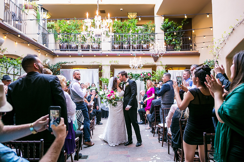 San juan capistrano outdoor wedding at plaza de magdelena bride form fitting gown with lace detail on bodice and thin straps with crystal belt and groom black notch lapel tuxedo with white dress shirt and black bow tie and white floral boutonniere kissing in the aisle after ceremony