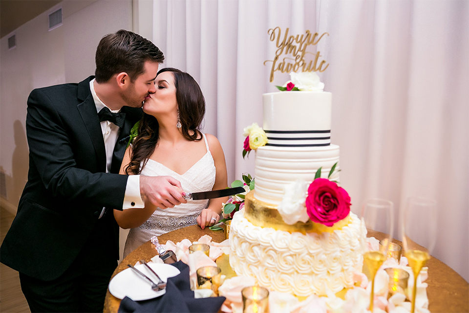 San juan capistrano outdoor wedding at plaza de magdelena bride form fitting gown with lace detail on bodice and thin straps with crystal belt and groom black notch lapel tuxedo with white dress shirt and black bow tie and white floral boutonniere kissing and cutting cake