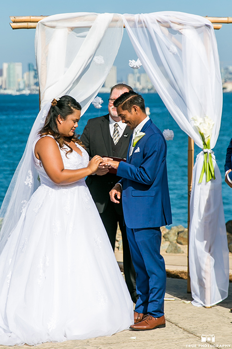 San diego outdoor wedding at bali hai bride ball gown with thin lace straps and a sweetheart neckline with lace and beading detail on bodice with long veil with groom cobalt blue notch lapel suit with white dress shirt and white vest with long white striped tie and pocket square with white floral boutonniere exchanging rings during ceremony