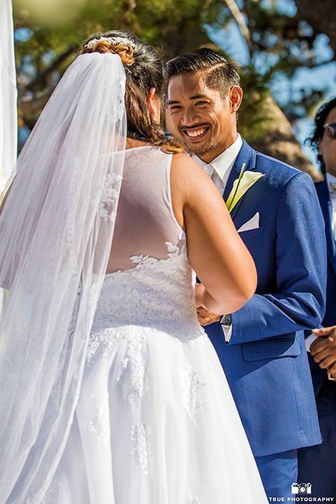 San diego outdoor wedding at bali hai bride ball gown with thin lace straps and a sweetheart neckline with lace and beading detail on bodice with long veil with groom cobalt blue notch lapel suit with white dress shirt and white vest with long white striped tie and pocket square with white floral boutonniere holding hands during ceremony