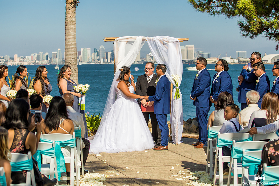 San diego outdoor wedding at bali hai bride ball gown with thin lace straps and a sweetheart neckline with lace and beading detail on bodice with long veil with groom cobalt blue notch lapel suit with white dress shirt and white vest with long white striped tie and pocket square with white floral boutonniere holding hands during ceremony