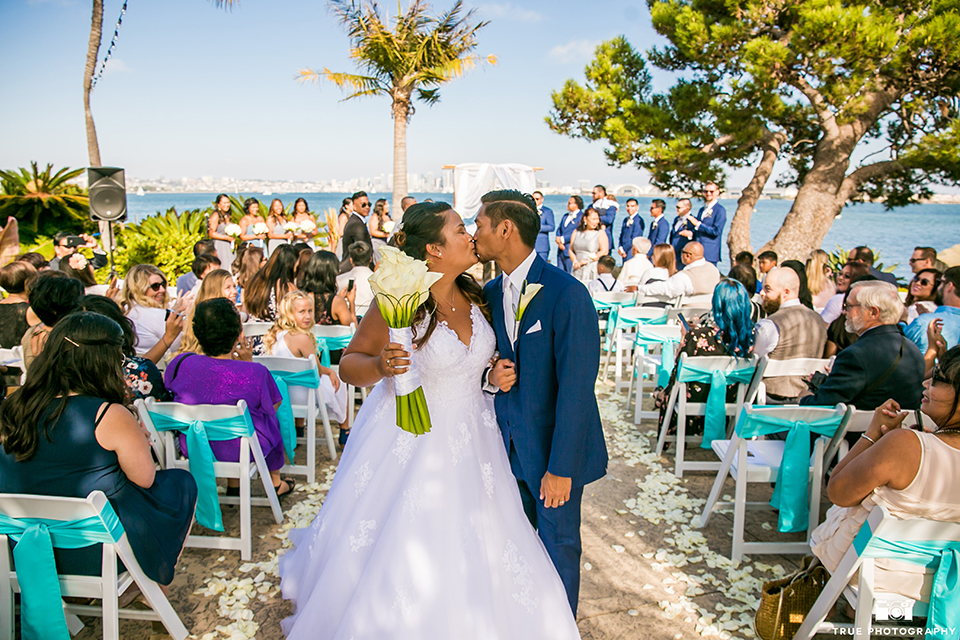 San diego outdoor wedding at bali hai bride ball gown with thin lace straps and a sweetheart neckline with lace and beading detail on bodice with long veil with groom cobalt blue notch lapel suit with white dress shirt and white vest with long white striped tie and pocket square with white floral boutonniere kissing at the end of the aisle after ceremony