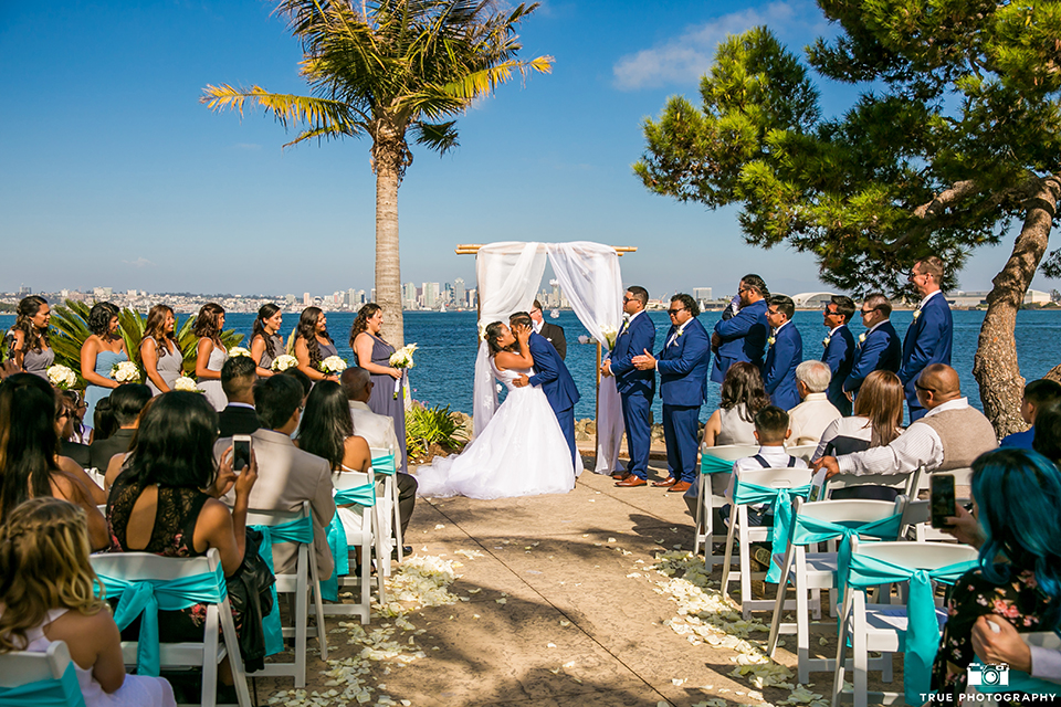 San diego outdoor wedding at bali hai bride ball gown with thin lace straps and a sweetheart neckline with lace and beading detail on bodice with long veil with groom cobalt blue notch lapel suit with white dress shirt and white vest with long white striped tie and pocket square with white floral boutonniere kissing during ceremony