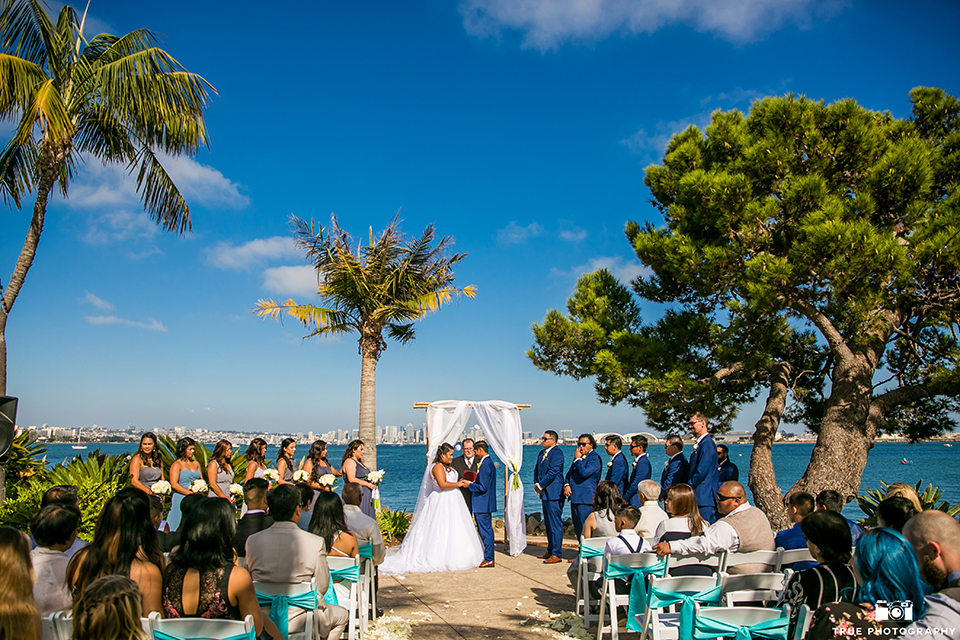 San diego outdoor wedding at bali hai bride ball gown with thin lace straps and a sweetheart neckline with lace and beading detail on bodice with long veil with groom cobalt blue notch lapel suit with white dress shirt and white vest with long white striped tie and pocket square with white floral boutonniere holding hands during ceremony