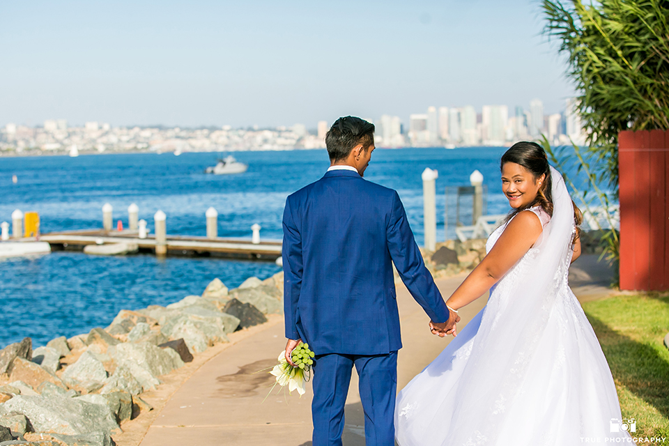 San diego outdoor wedding at bali hai bride ball gown with thin lace straps and a sweetheart neckline with lace and beading detail on bodice with long veil with groom cobalt blue notch lapel suit with white dress shirt and white vest with long white striped tie and pocket square with white floral boutonniere walking and holding hands