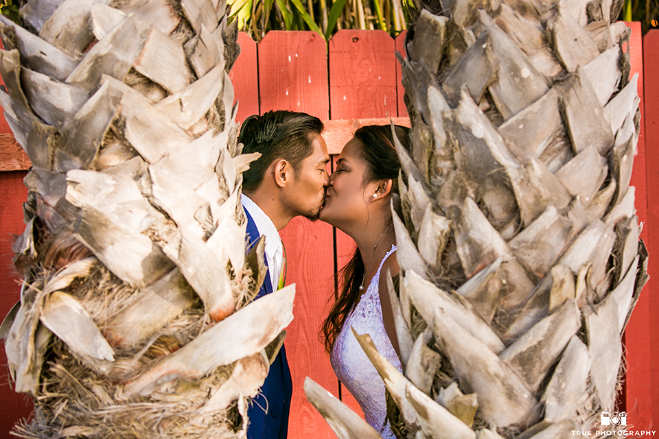 San diego outdoor wedding at bali hai bride ball gown with thin lace straps and a sweetheart neckline with lace and beading detail on bodice with long veil with groom cobalt blue notch lapel suit with white dress shirt and white vest with long white striped tie and pocket square with white floral boutonniere kissing behind tree