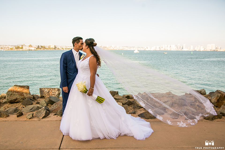 San diego outdoor wedding at bali hai bride ball gown with thin lace straps and a sweetheart neckline with lace and beading detail on bodice with long veil with groom cobalt blue notch lapel suit with white dress shirt and white vest with long white striped tie and pocket square with white floral boutonniere standing and kissing