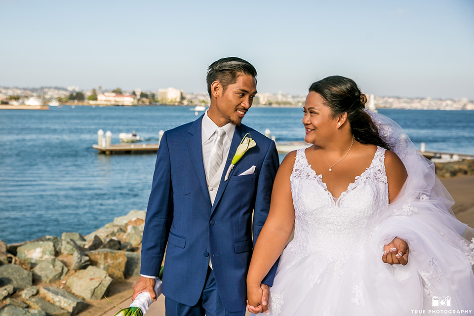 San diego outdoor wedding at bali hai bride ball gown with thin lace straps and a sweetheart neckline with lace and beading detail on bodice with long veil with groom cobalt blue notch lapel suit with white dress shirt and white vest with long white striped tie and pocket square with white floral boutonniere walking and holding hands