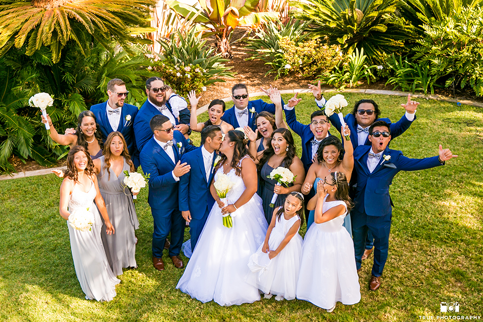 San diego outdoor wedding at bali hai bride ball gown with thin lace straps and a sweetheart neckline with lace and beading detail on bodice with long veil with groom cobalt blue notch lapel suit with white dress shirt and white vest with long white striped tie and pocket square with white floral boutonniere kissing with wedding party cheering