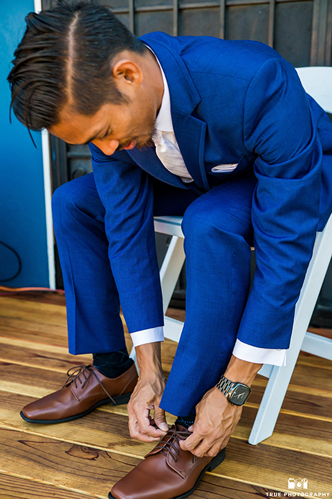 San diego outdoor wedding shoot at bali hai groom cobalt blue notch lapel suit with a white dress shirt and white vest with a long white striped tie and pocket square putting on shoes getting ready before ceremony