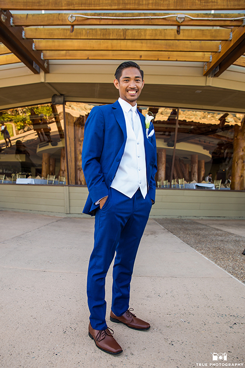 San diego outdoor wedding shoot at bali hai groom cobalt blue notch lapel suit with a white dress shirt and white vest with a long white striped tie and pocket square with white floral boutonniere standing with hands in pockets